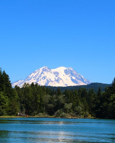 Snow-capped mountain with forested shoreline and blue lake in foreground under clear sky