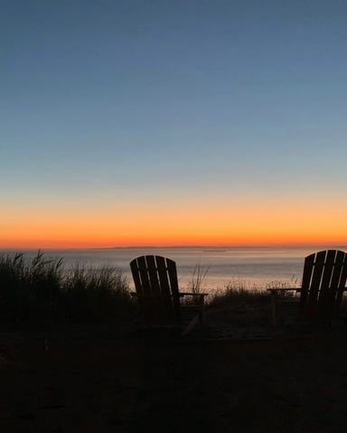 Two Adirondack chairs silhouetted on beach at sunset overlooking calm water