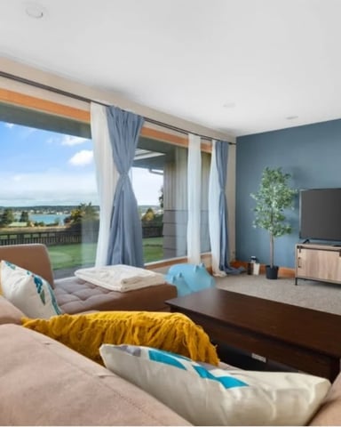 Modern living room with beige sectional sofa, TV, and large windows overlooking lake and green landscape