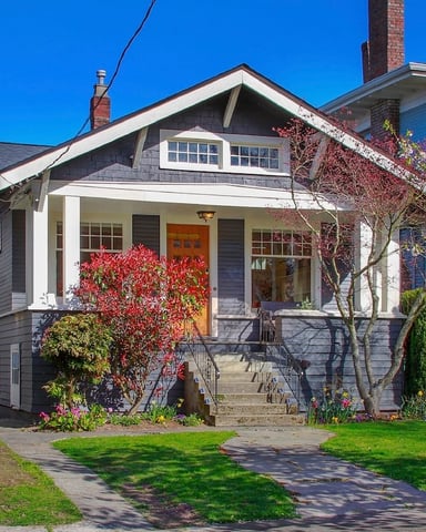 Craftsman-style house with white trim, dark siding, and orange door, surrounded by blooming spring trees and manicured lawn.