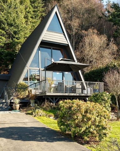 Modern A-frame cabin with dark exterior and large windows, elevated deck, surrounded by evergreen forest and landscaped grounds.