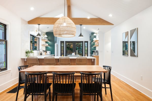 Modern open-plan kitchen and dining room with wooden beams, woven pendant light, green cabinetry, and wood dining table with black chairs