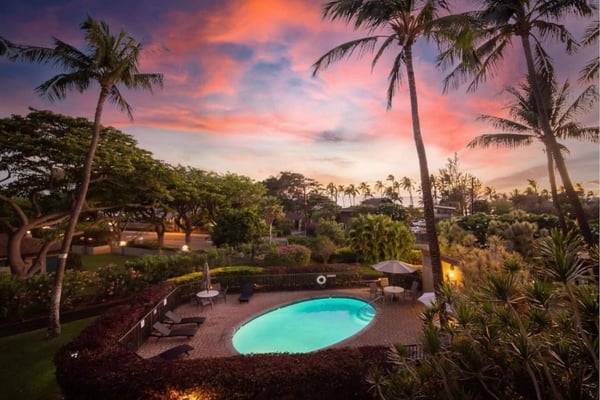 Illuminated pool surrounded by palm trees and tropical landscaping at sunset with colorful sky