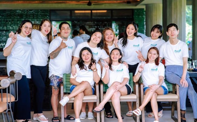 Group of twelve people wearing matching white t-shirts posing together indoors at a casual venue with green plant wall backdrop