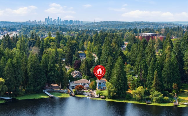 Waterfront home with red location pin marker, surrounded by evergreen trees, with city skyline visible in distance